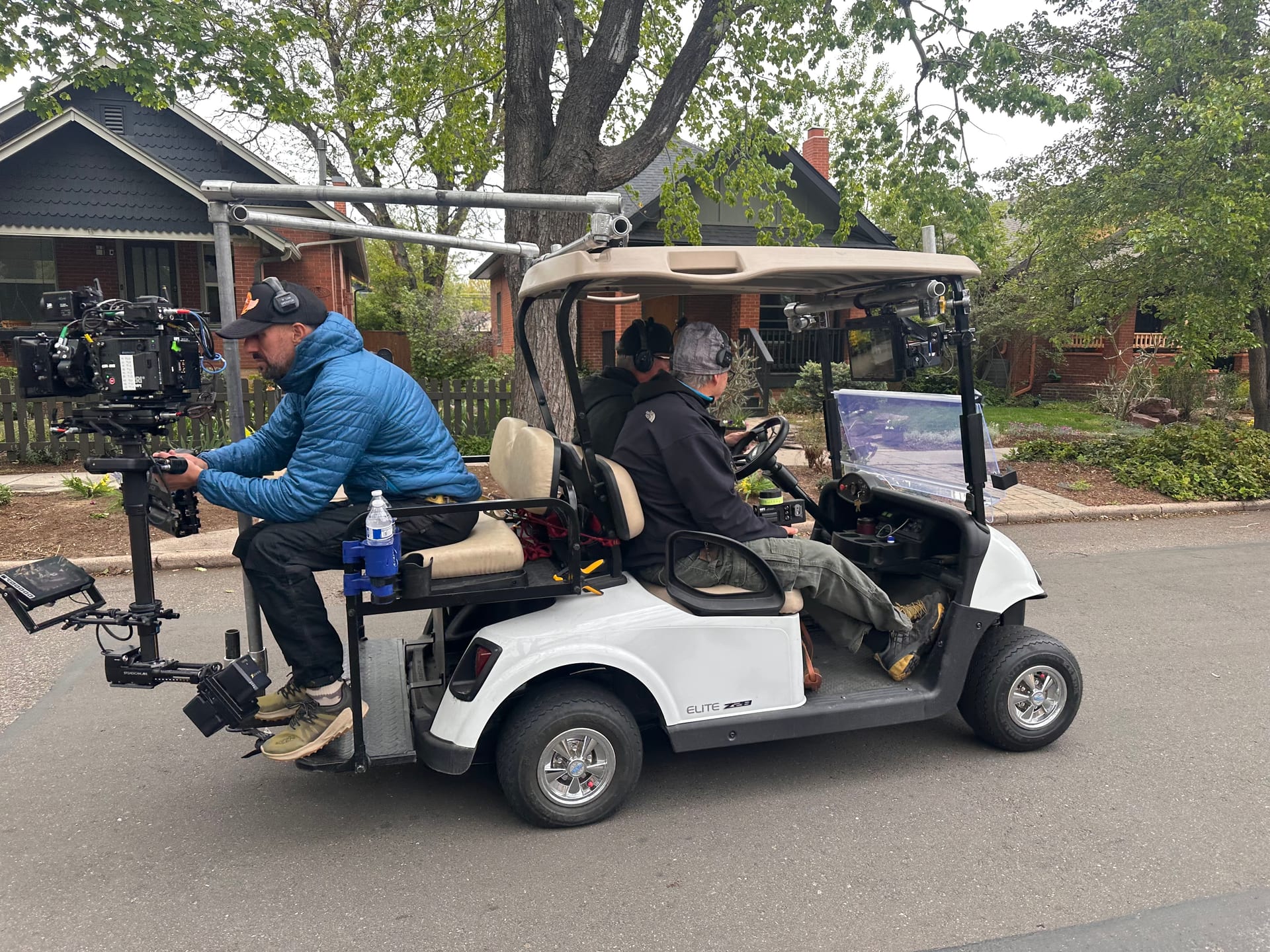 Camera operator seated at the rear of the moving camera car, filming with an ARRI cinema camera on a stabilizer rig while the cart travels along a quiet Colorado residential road lined with trees and houses.